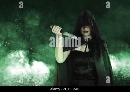 Donna strega asiatica con mantello nero che tiene in piedi un coltello con fuoco mistico su sfondo nero Foto Stock