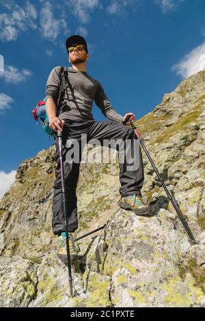 Portatore di un elegante viaggiatore hipster con una barba e uno zaino in occhiali da sole e un cappello con bastoni da trekking in piedi su una roccia Foto Stock