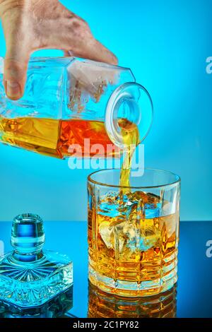 Versare decantere di cristallo quadrato con nastro scotch whiskey o brandy in un vetro rotondo di cristallo con ghiaccio su uno sfondo blu gradiente con riflessione. Co Foto Stock
