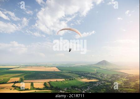 Il parapendio professionista in una tuta da cocco vola in alto sopra il terreno contro il cielo e i campi con le montagne Foto Stock