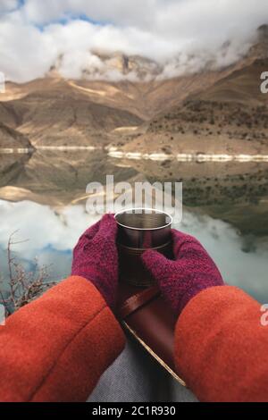 Una vista in prima persona delle mani delle donne in camici e mittenne stanno tenendo una tazza di metallo con tè o caffè e un diario tascabile in pelle Foto Stock