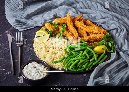 bastoncini di pesce, barrette di filetto di pesce impanate e fritte servite su un piatto nero con couscous e fagioli verdi al vapore su un tavolo di cemento con tartaro Foto Stock