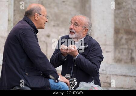 Due italiani parlano tra loro e fanno gesti tipici italiani sulla piazza del Popolo di Ascoli Piceno, Italia
