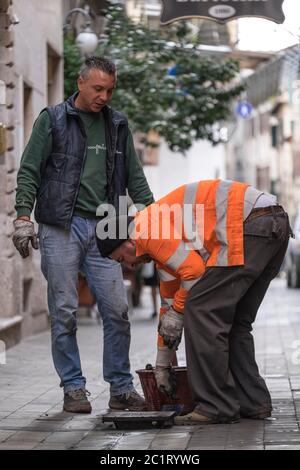 Due operai aprono un tombino fognario non protetto in una strada della città di Ascoli Picena, Italia Foto Stock