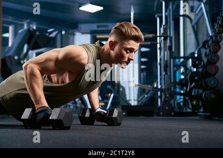 Raccolto di addestramento dell'uomo caucasico muscolare sul pavimento usando i manubri nel randello di sport. Primo piano di giovani sportivi che costruiscono muscoli, facendo spinta in su in palestra vuota. Concetto di sport, bodybuilding. Foto Stock