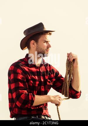 Concetto di agricoltura. Uomo bello in hat e stile rustico vestito. Tenere il ranch. La vita al ranch. Cowboy con lazo corda sullo sfondo del cielo. Riflessivo agricoltore pensando di business. Ranch lavoratore. Fattoria di eco. Foto Stock