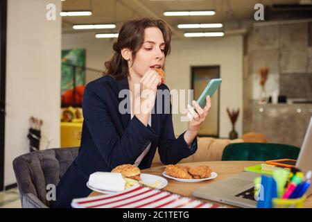 Donna che mangia hamburger seduto in un ufficio moderno sul posto di lavoro. Foto Stock