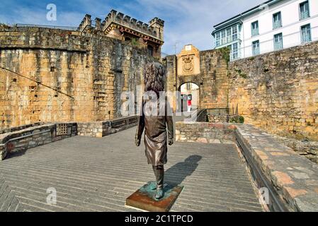 Statua in bronzo e donna con pullover rosso che attraversa il portale medievale (Hondurribia, Basaue Country) Foto Stock