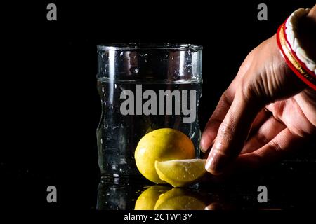 Un bicchiere d'acqua posto su una superficie riflettente su uno sfondo scuro con limone intatto e tagliato posto di fronte ad esso e tenuto da una mano Foto Stock