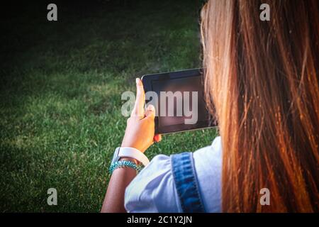 Una giovane donna si rilassa sull'erba del parco e fa una videochiamata sul suo tablet sopra la spalla shot Foto Stock