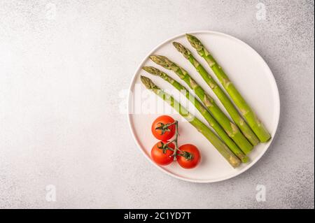 Asparagi freschi e pomodori ciliegini su un piatto bianco su uno sfondo chiaro con vista dall'alto dello spazio per la copia Foto Stock