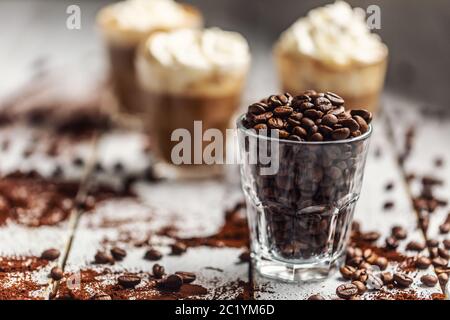 Chicchi di caffè in una tazza di vetro e sullo sfondo una tazza con caffè e panna montata Foto Stock