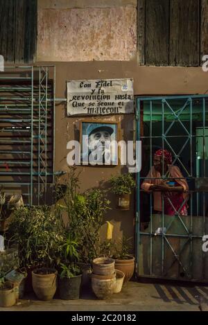 Donna che guarda attraverso i bar della sua porta d'ingresso accanto ad una foto di Fidel Castro a l'Avana Centro, un quartiere di classe operaia, l'Avana, Cuba Foto Stock