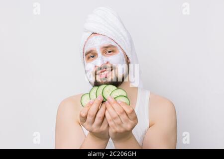 Primo piano ritratto di felice divertente uomo bearded in asciugamano bianco con maschera facciale in argilla per la cura della pelle, avendo divertimento tenendo le fette di cetriolo isolate sopra Foto Stock