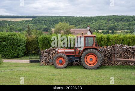 Trattore rosso parcheggiato in un giardino di fronte a una pila di legno tagliato. Paesaggio di campagna. Foto Stock