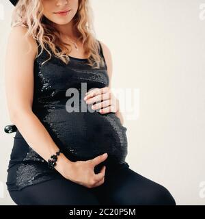 Raccolto di donna bionda incinta in elegante top brillante abbracciando il suo pancino. Foto Stock