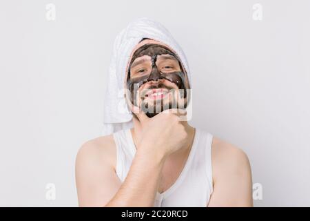 Felice uomo orgoglioso con maschera nera sul viso. Foto di bell'uomo bearded che riceve trattamenti termali, che si posa a macchina fotografica su isolato sfondo bianco Foto Stock