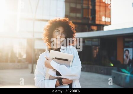 Adorabile imprenditore dai capelli ricci con occhiali che si posano all'esterno in una camicia bianca che abbraccia un computer portatile Foto Stock