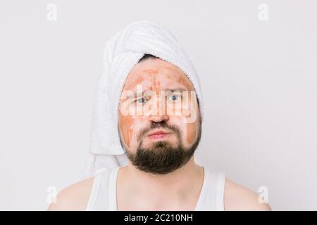 Divertente uomo bearded bello con asciugamano bianco sulla testa, con una maschera marrone fango sul viso, guardando la macchina fotografica su sfondo bianco. Uomo con volto espressivo Foto Stock