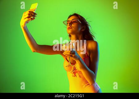 Selfie. Bel ritratto di giovane ragazza su sfondo studio gradiente in neon. Donna in tuta alla moda con cocktail. Espressione facciale, estate, fine settimana, bellezza, concetto di resort. Vacanze. Foto Stock
