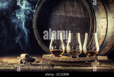 Bicchieri per degustazione di whisky Glencairn su una porzione di legno, con un barile di whisky sullo sfondo scuro e un sigaro affumicante accanto ad esso Foto Stock