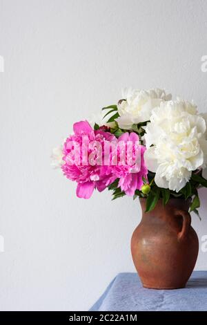 Bel bouquet di peonie in una pentola di argilla si erge su un tavolo con una tovaglia di lino blu Foto Stock