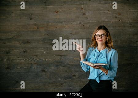 Bella donna tiene un libro e che mostra lo spazio di copia. Vestito in blu camicetta, in occhiali. Grigio sfondo di legno Foto Stock