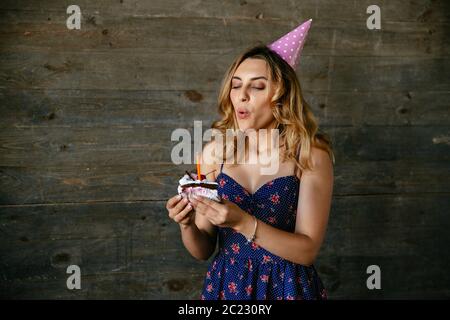 Affascinante ragazza celebrare la festa di compleanno, soffiando fuori la candela sulla crema torta al cioccolato, festosa che indossa un cappello e abiti colorati. Foto Stock