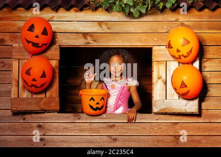 Ragazza dei capelli ricci sorridente piccola in costume di Halloween con secchio per le caramelle sguardo dalla finestra del playhouse Foto Stock