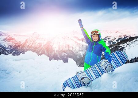 Felice ragazzo stese con snowboard in ascensore mostra mano grande sorriso sopra montagna alta cima sullo sfondo Foto Stock