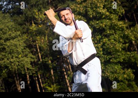 Il maestro delle arti marziali nel kimono bianco sta praticando con nunchaku all'aperto nella natura. Foto Stock