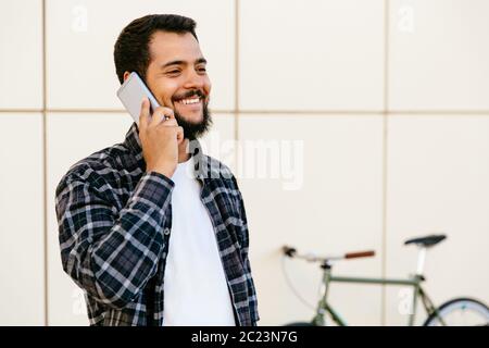 Allegro elegante giovane uomo barbuto comunicanti tramite un telefono cellulare avente una piacevole conversazione, permanente, mentre all'aperto vicino alla bicicletta. Foto Stock