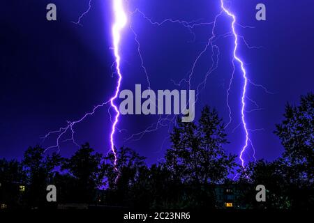 lampi luminosi di fulmini durante temporali notturni, sagome di alberi e case contro il cielo blu scuro Foto Stock