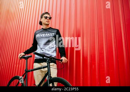 Uomo premuroso in berretto alla moda e occhiali da sole che tengono una bicicletta, in piedi vicino al muro rosso, all'aperto. Foto Stock