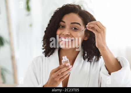 Sorridente Afro Donna applicando idratante viso Serum sulla sua bella pelle Foto Stock