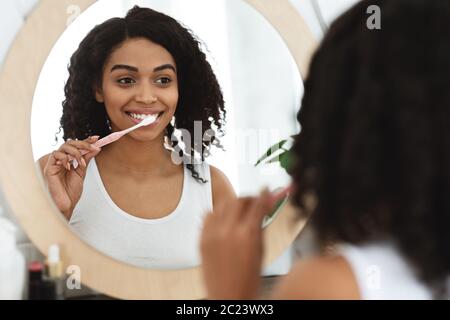 Dental Care quotidiana. Donna africana sorridente spazzolando i denti vicino a Specchio in bagno Foto Stock