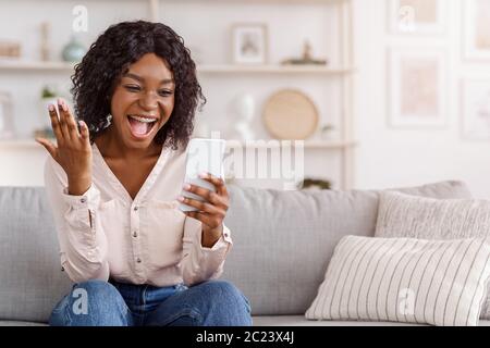 La ragazza nera molto gioiosa che festeggia il successo con lo smartphone a casa, ha ricevuto buone notizie Foto Stock