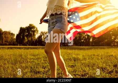 Giornata dell'indipendenza USA. Ragazza in pantaloncini con bandiera americana corre sull'erba in un parco estivo al tramonto. Foto Stock