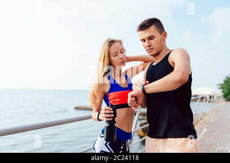 Muscoloso uomo bello e sportivi ragazza controllo una volta su smart orologi, dopo l'esecuzione di allenamento sul quay, vicino al mare. Sport concept. Foto Stock