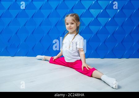 Una bambina con i pigtail in una T-shirt bianca su uno sfondo blu si siede in spago sul pavimento e sorride. Emozioni dei bambini occupazione spot, divertimento Foto Stock