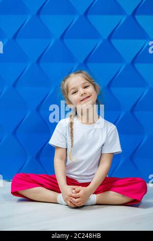 Bambina con cavetti in una T-shirt bianca su sfondo blu. Si siede in una posizione di loto sul pavimento e sorride e le ondeggia le mani. Foto Stock