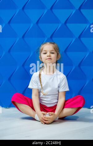 Bambina con cavetti in una T-shirt bianca su sfondo blu. Si siede in una posizione di loto sul pavimento e sorride e le ondeggia le mani. Foto Stock