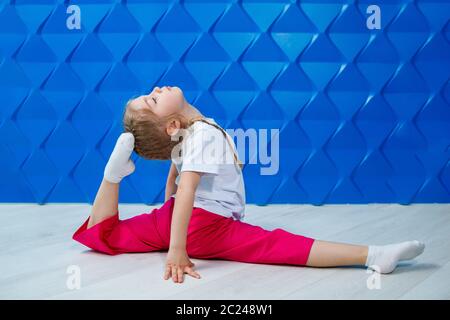 Una bambina con i pigtail in una T-shirt bianca su uno sfondo blu si siede in spago sul pavimento e sorride. Emozioni dei bambini occupazione spot, divertimento Foto Stock