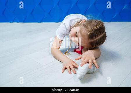 Una bambina con i pigtail in una T-shirt bianca su uno sfondo blu si siede in spago sul pavimento e sorride. Emozioni dei bambini occupazione spot, divertimento Foto Stock