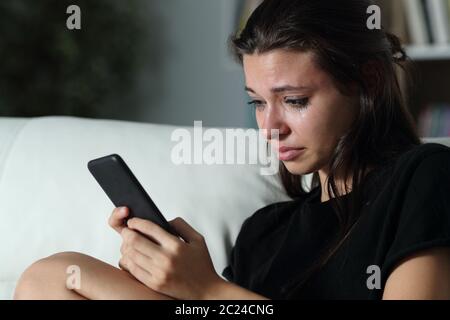 Triste ragazza adolescente controllo smart phone cyber bullismo messaggi seduto su un divano nel buio a casa Foto Stock