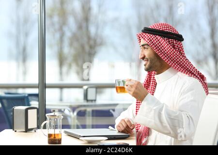 Vista laterale verticale di un felice uomo arabo che guarda lontano attraverso una finestra tenendo una tazza da tè seduti in un caffè Foto Stock