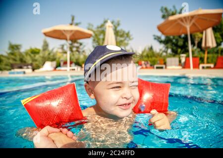 Un piccolo ragazzo impara a nuotare in piscina in estate con il sostegno di suo padre le mani Foto Stock