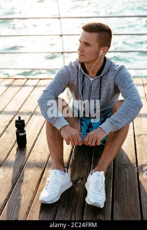 Giovani pensieroso sportsman ascoltando musica in cuffia, guardando lontano, seduta sul molo, con bottiglia di acqua. Vestito in abbigliamento sportivo. Vicino al mare. Foto Stock