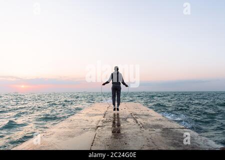 Lunghezza piena vista del giovane sportivo del salto con la corda, allenamento sul molo, vicino al mare. Vista dal retro. Foto Stock
