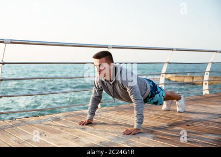 Foto all'aperto di un uomo sportivo nelle cuffie facendo push-up esercizi, durante l'allenamento sul quay, vicino all'oceano. Foto Stock